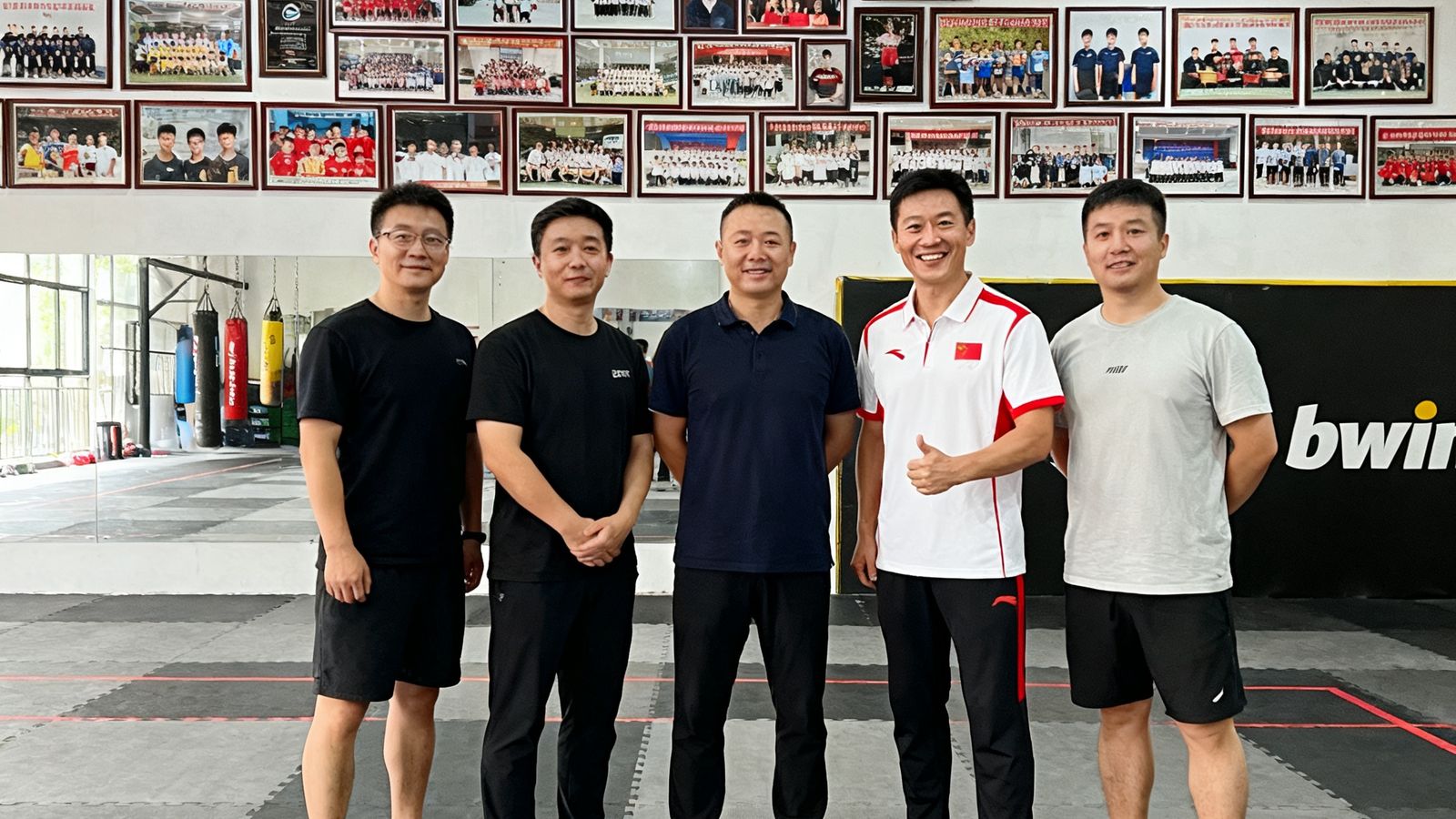 bwin创始团队五位核心成员站在训练馆中央，背景墙上密密麻麻挂满了历届学员获得的赛事奖牌和毕业合影照片，其中一位身穿国家队队服的创始人正对着镜头微笑，地面铺设专业运动地胶